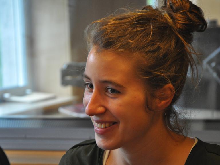 Eine junge Frau mit lockigen Haaren sitzt lächelnd in einem Tonstudio.