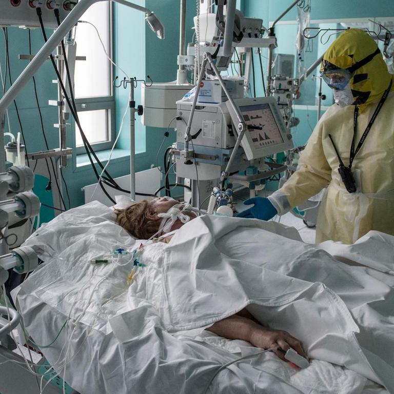 Der Arzt in voller Schutzkleidung steht an der Bettseite der Patientin, die unter einer weißen Decke liegt. Um sie herum Geräte, Kabel und Schläuche.