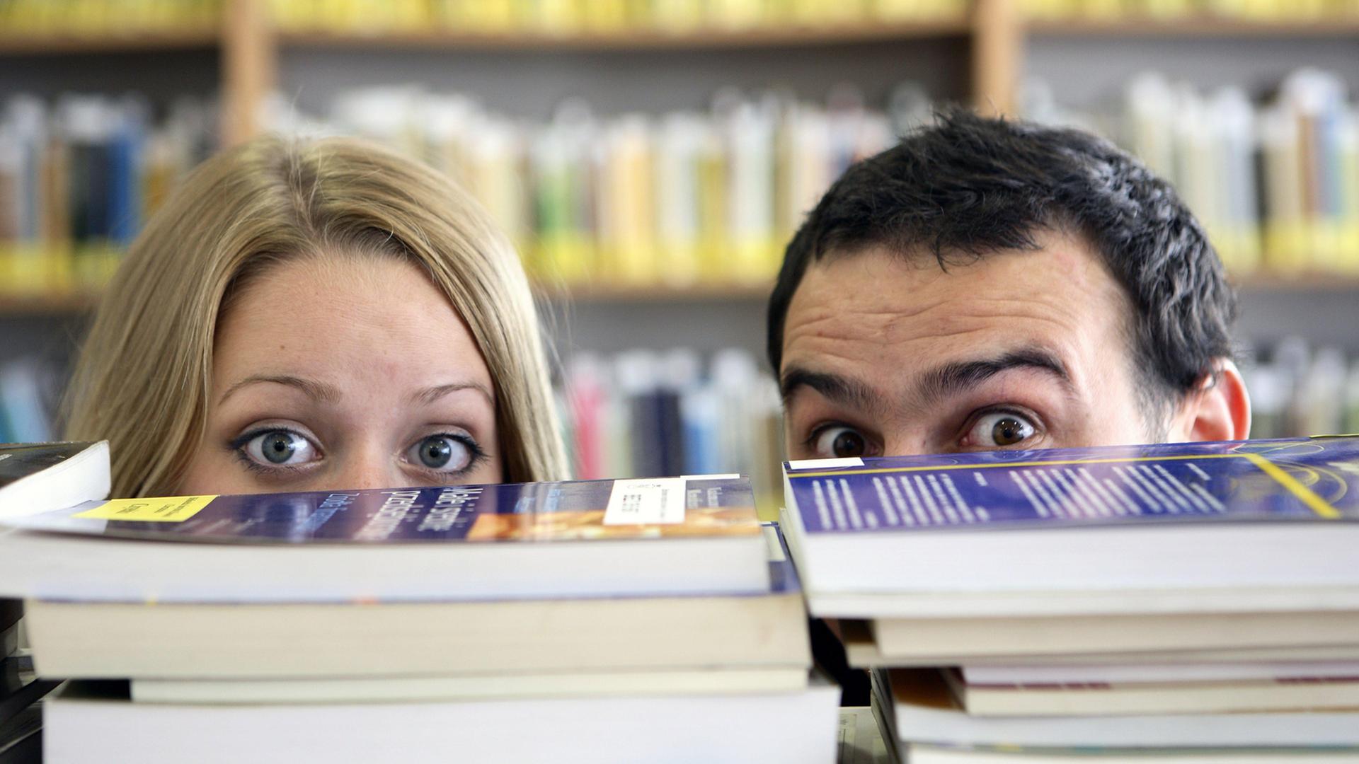 Ein Man und eine Frau verstecken sich hinter einem Stapel Bücher. Von den Gesichtern sind nur die großen offenen Augen und der Haaransatz zu sehen.