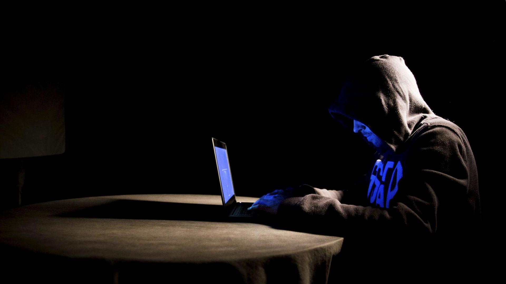 Mann mit Kapuze über dem Kopf sitzt bei sehr schwacher Beleuchtung an einem Tisch. Auf dem Tisch steht ein Laptop.