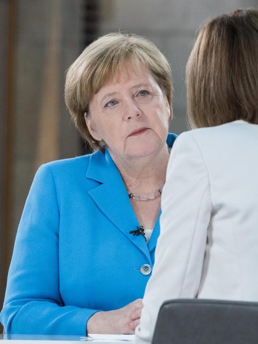 Die Interviewerin im weißen Jackett sieht man von hinten an einem Tisch sitzen. Ihr gegenüber sitzt Merkel im blauen Jackett und hört aufmerksam zu.
