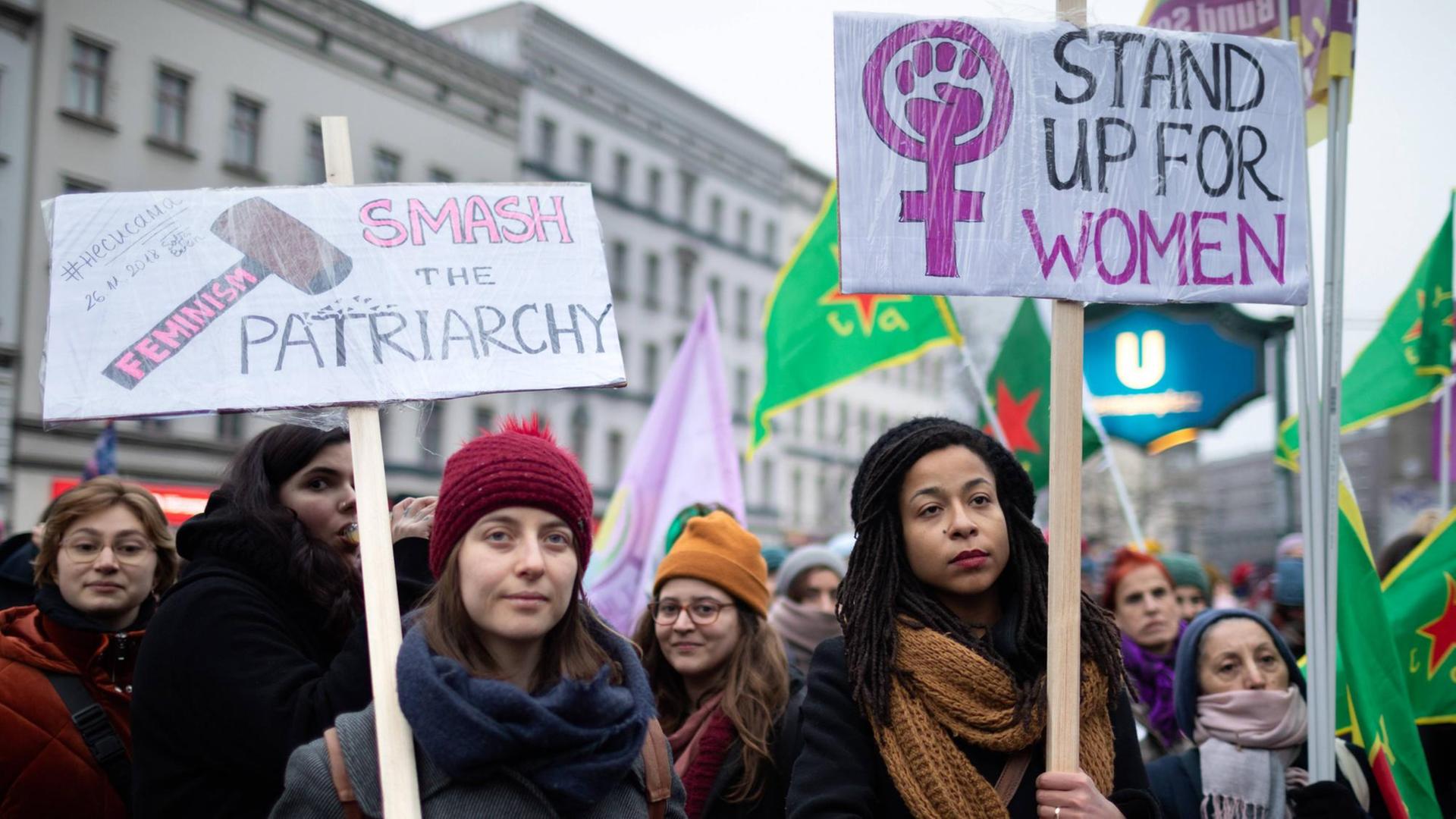 Zwei Frauen halten Plakate, auf denen steht "Smash the Patriarchy" und "Stand up for Women"