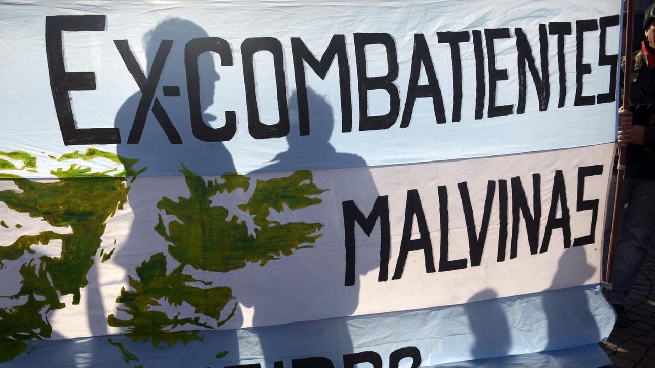 Sie sehen die Schatten von zwei Menschen hinter einer argentinischen Flagge, auf der steht: Ex-Kämpfer Malvinas. Malvinas ist der argentinische Name für die Falkland-Inseln.