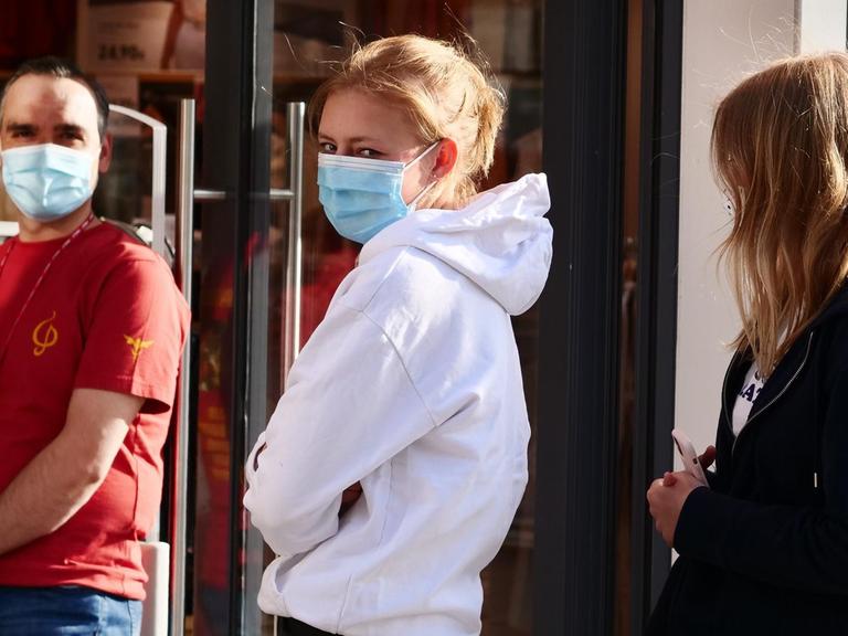 Zwei junge Frauen stehen vor einem Bekleidungsgeschaeft Schlange mit Abstand zueinander und mit Masken gegen das Coronavirus.