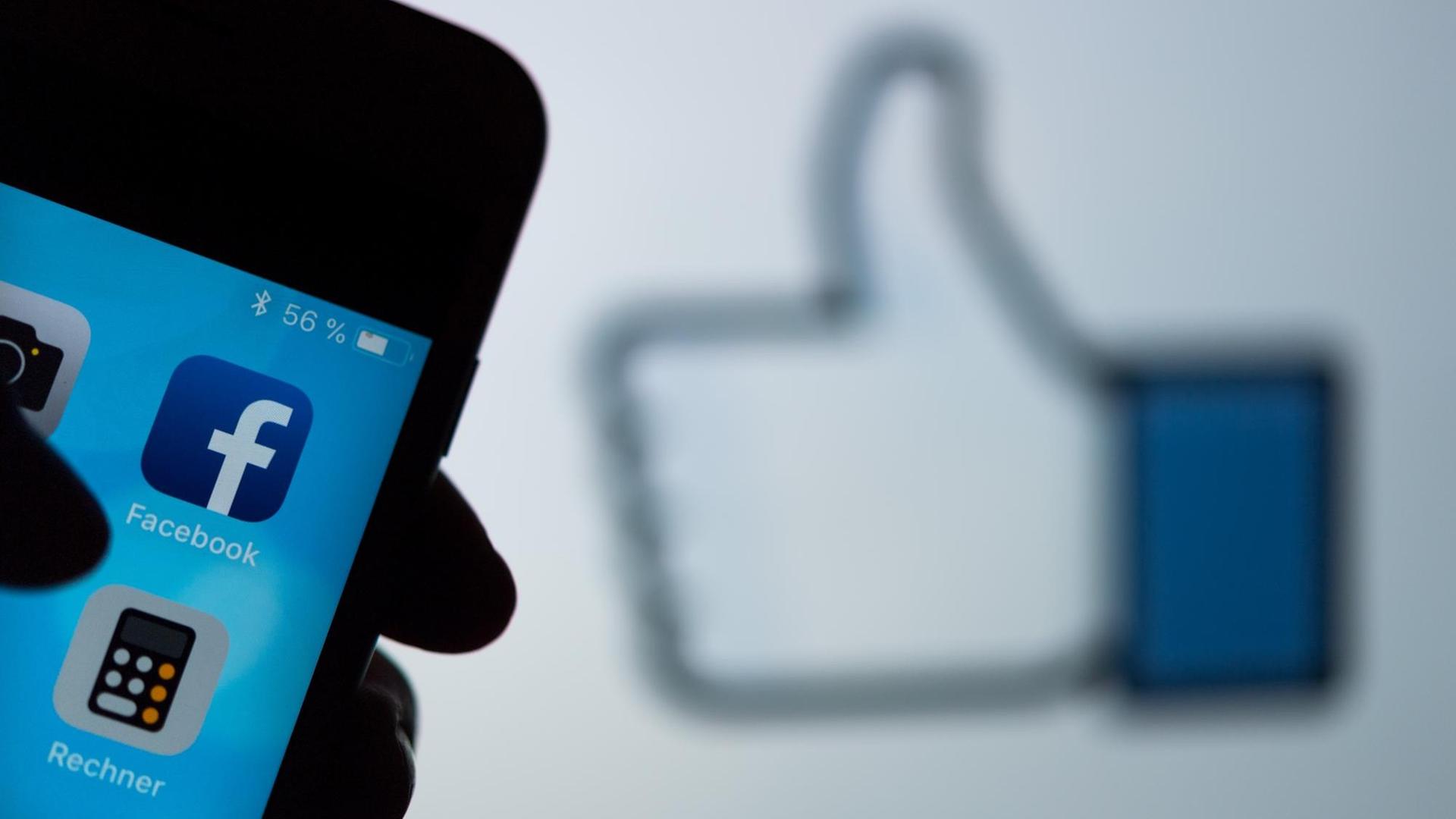 Das Logo des sozialen Netzwerks Facebook ist auf dem Display eines Smartphones zu sehen. Im Hintergrund wird auf einem Bildschirm ein das Zeichen für "Gefällt mir" (Daumen hoch) angezeigt.
