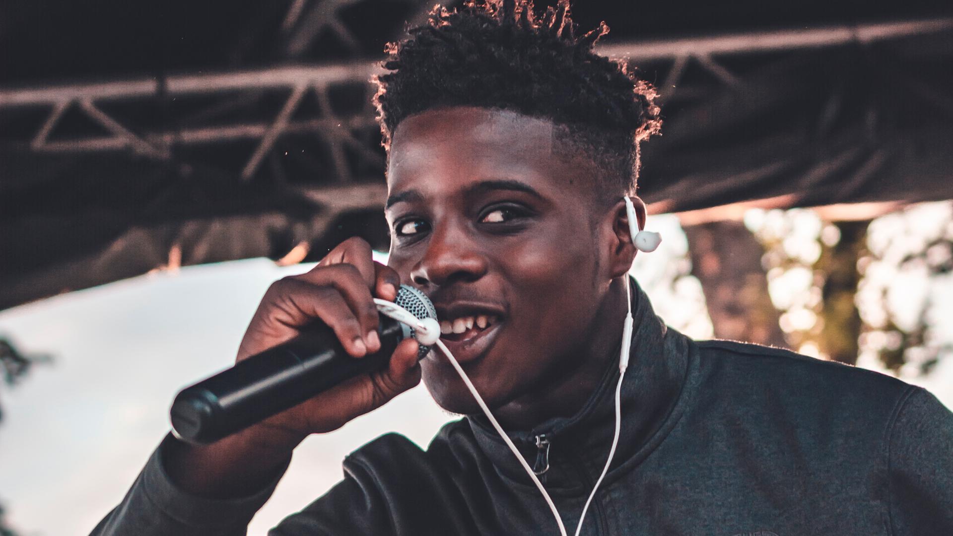 Ein junger schwarzer Mann, der sehr optimistisch aussieht, hält ein Mikrofon in der Hand und singt.