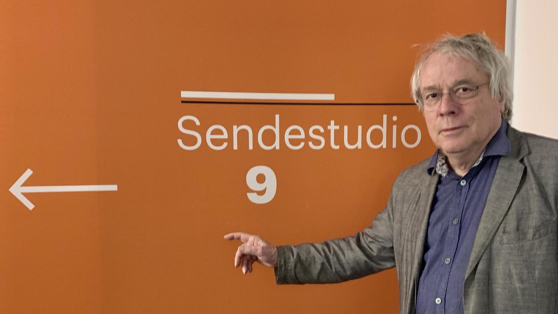 Der Journalist Thomas Nehls deutet im Funkhaus von Deutschlandfunk Kultur in Berlin auf einen Wegweiser "Sendestudio 9" auf einer orangefarbenen Wand.