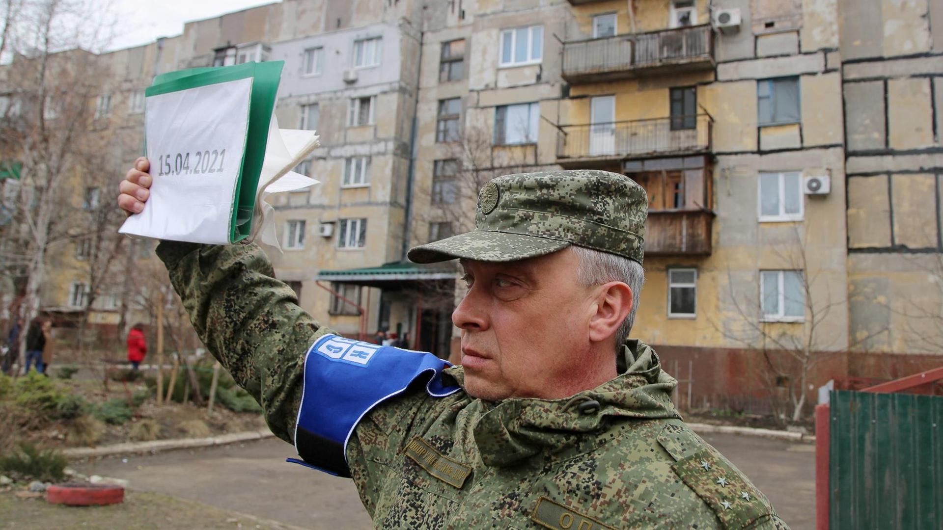 Ein Mann in Tarnfarben-Uniform und blauer Armbinde hält ein Schild mit dem datum '15.04.2021' vor einer Reihe Wohnhäuser hoch.