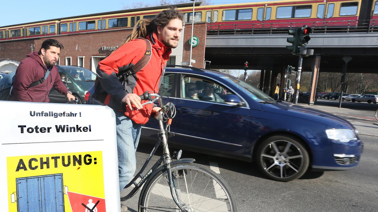Fahrradfahrer fahren auf einem Fahrradweg an einer Kreuzung in Berlin geradeaus, während ein Auto rechts abbiegen will, davor weist ein Schild auf den Toten Winkel als Unfallgefahr hin.