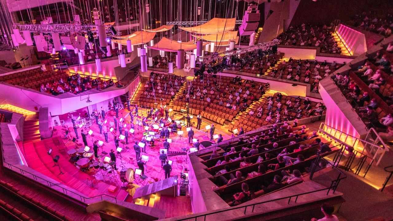 Blick von einem oberen Rang in den Zuschauersaal der Philharmonie mit der zentralen Bühne, der in gelbes und lilafarbenes Licht getaucht ist.