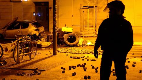 Die Silhouette eines Polizisten in Kampfmonitur in der gelblich beleuchteten Straße. Im Hintergrund ein ausgebranntes Auto und viele Pflasterssteine.