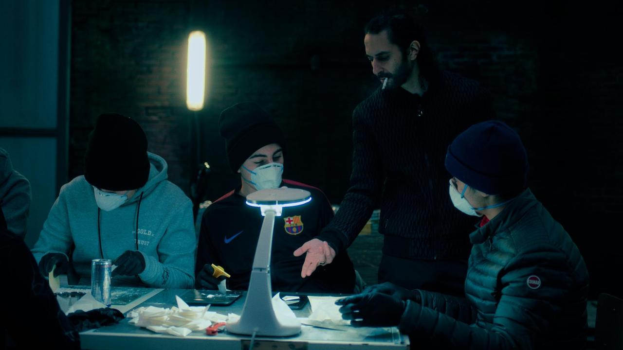 Die Filmszene aus der Serie "Mocro Maffia" zeigt eine Drogenlabor. In einem spärlich beleuchteten Raum sitzen jungen Männer mit Gesichtsmasken, ein älterer Mann gibt ihnen Anweisungen.