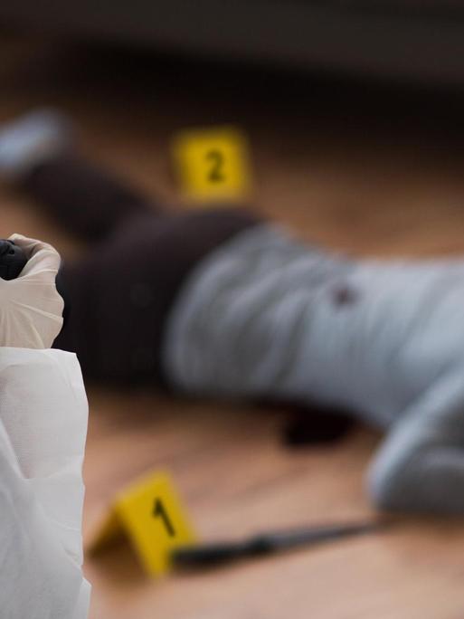 Nachgestellte Tatortszene: Kriminalist im weißen Schutzanzug und mit Kamera, der einen toten weiblichen Körper am Tatort fotografiert.