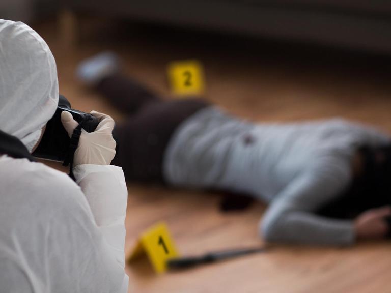 Nachgestellte Tatortszene: Kriminalist im weißen Schutzanzug und mit Kamera, der einen toten weiblichen Körper am Tatort fotografiert.