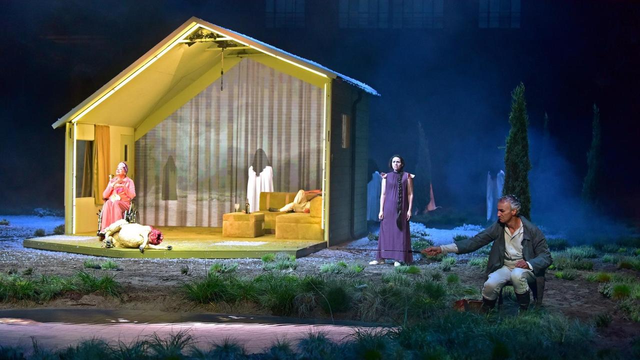 Eine Holzhütte mit offener Front steht in einer Halle als Bühnenbild, zwei Personen befinden sich in der Hütte, zwei rechts neben ihr. Außerhalb der Hütte leuchtet dunkelblaues Licht als Nachtstimmung.