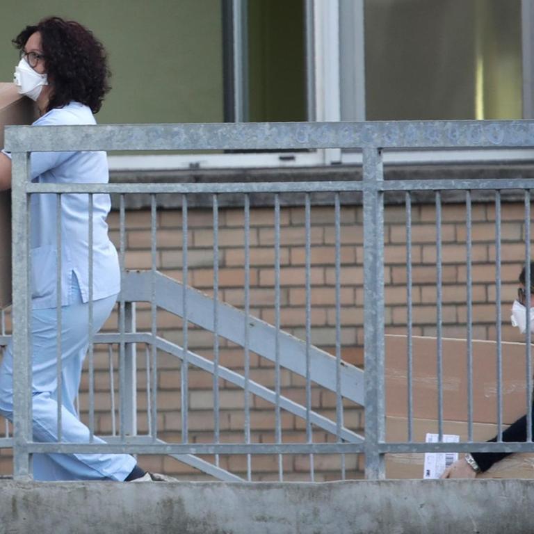 Zwei Frauen mit Mundschutz und Krankenhausuniform gehen vor einer Backstein-Außenfassade eine Treppe aus einem Untergeschoß hoch und tragen Kartons.