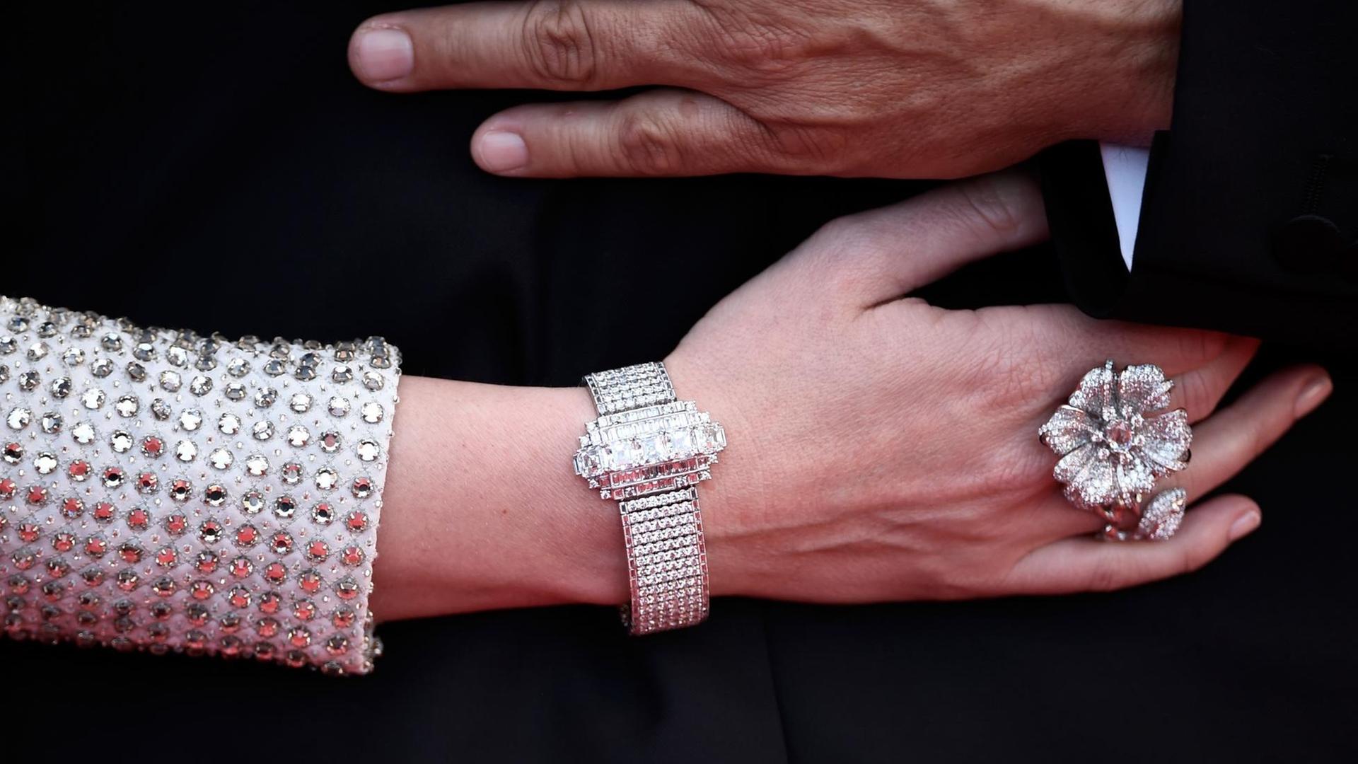 Eine Frauenhand mit grossem Diamantenring und-armband und silber glitzerndem Ärmel. Darüber eine Männerhand.