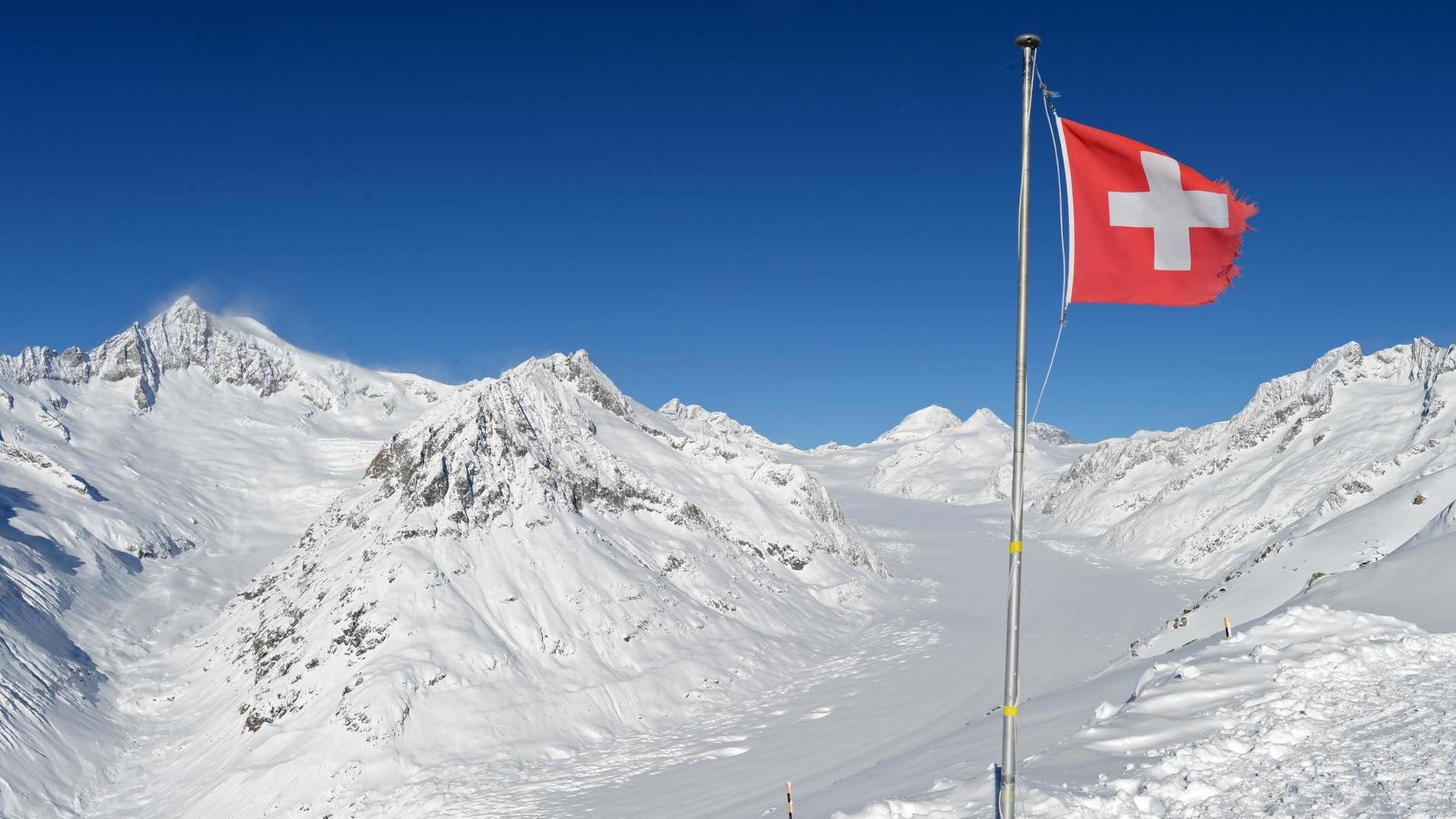 Die Schweizer Nationalfahne weht bei strahlendem Sonnenschein nahe dem Eggishorn bei Fiesch (Wallis) in der Schweiz, im Hintergrund ist der Große Aletsch.
