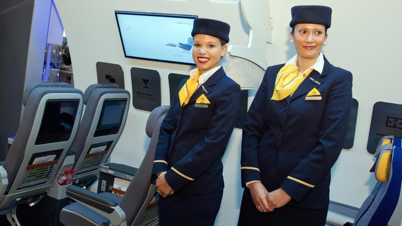 Die beiden Stewardessen stehen im Nachbau eines Lufthansa-Innenraums. Sie tragen blaue Lufthansa-Uniformen mit gelben Halstüchern und blaue Mützen. Beide lächeln in die Kameras. Die beiden Stewardessen stehen im Nachbau eines Lufthansa-Innenraums. Sie tragen blaue Lufthansa-Uniformen mit gelben Halstüchern und blaue Mützen. Beide lächeln in die Kameras.