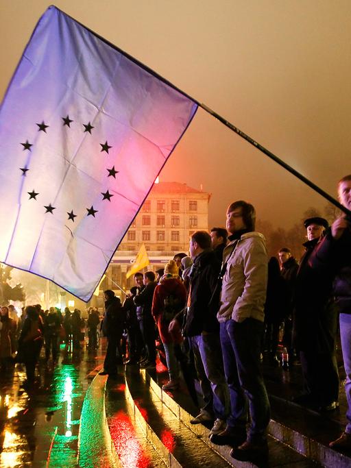 Mehrere Demonstranten stehen am Rand einer Straße, einer hält eine große Flagge mit dem Logo der Europäischen Union.