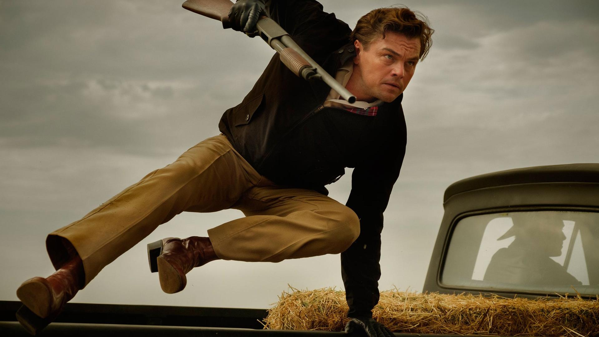 Das Bild zeigt den Schauspieler Leoonardo DiCaprio beim Sprung über ein Auto mit einem Gewehr in der Hand.