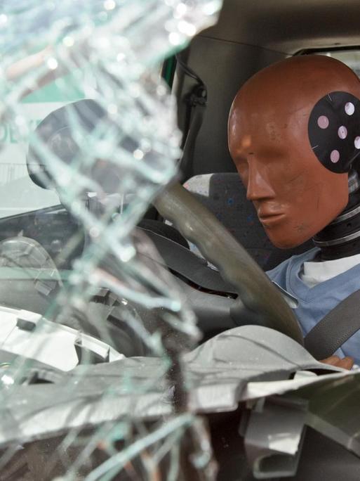 Eine Crashtestpuppe sitzt nach einer Frontalkollision mit einem anderen PKW hinter zerborstetem Glas und Metall in einem Versuchsfahrzeug.