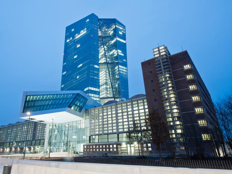 Außenansicht des EZB-Neubaus in Frankfurt in der Abenddämmerung. Hinter einem langgestreckten Gebäude erhebt sich ein gewundener Turm.