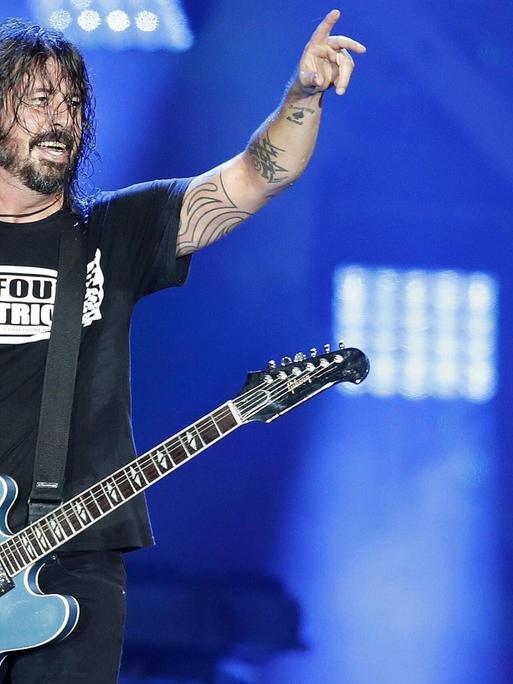 Ein Gitarrist mit langen, schwarzen, verschwitzten Haaren steht auf einer Bühne vor blauem Hintergrund, seine rechte Hand liegt auf den Saiten seiner Gitarre, die linke hält er auf Kopfhöhe in die Luft.