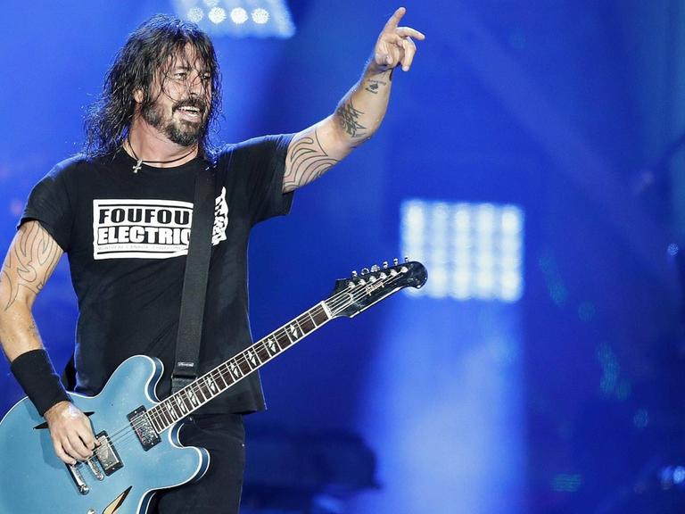 Ein Gitarrist mit langen, schwarzen, verschwitzten Haaren steht auf einer Bühne vor blauem Hintergrund, seine rechte Hand liegt auf den Saiten seiner Gitarre, die linke hält er auf Kopfhöhe in die Luft.