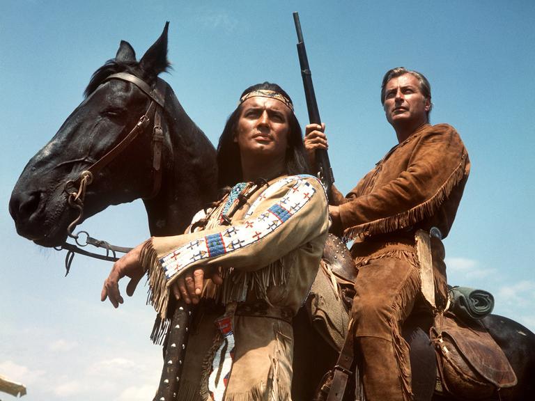Pierre Brice (l) als Apachen-Häuptling Winnetou und Lex Barker als sein Blutsbruder Old Shatterhand in einer Szene des Karl-May-Films "Im Tal des Todes". Lex Barker sitzt auf einem Pferd mit einem Gewehr in der Hand. Beide schauen in die Ferne.