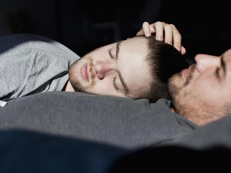 Ein schwules Paar liegt mit geschlossenen Augen im Bett und die Sonne scheint ihnen ins Gesicht.