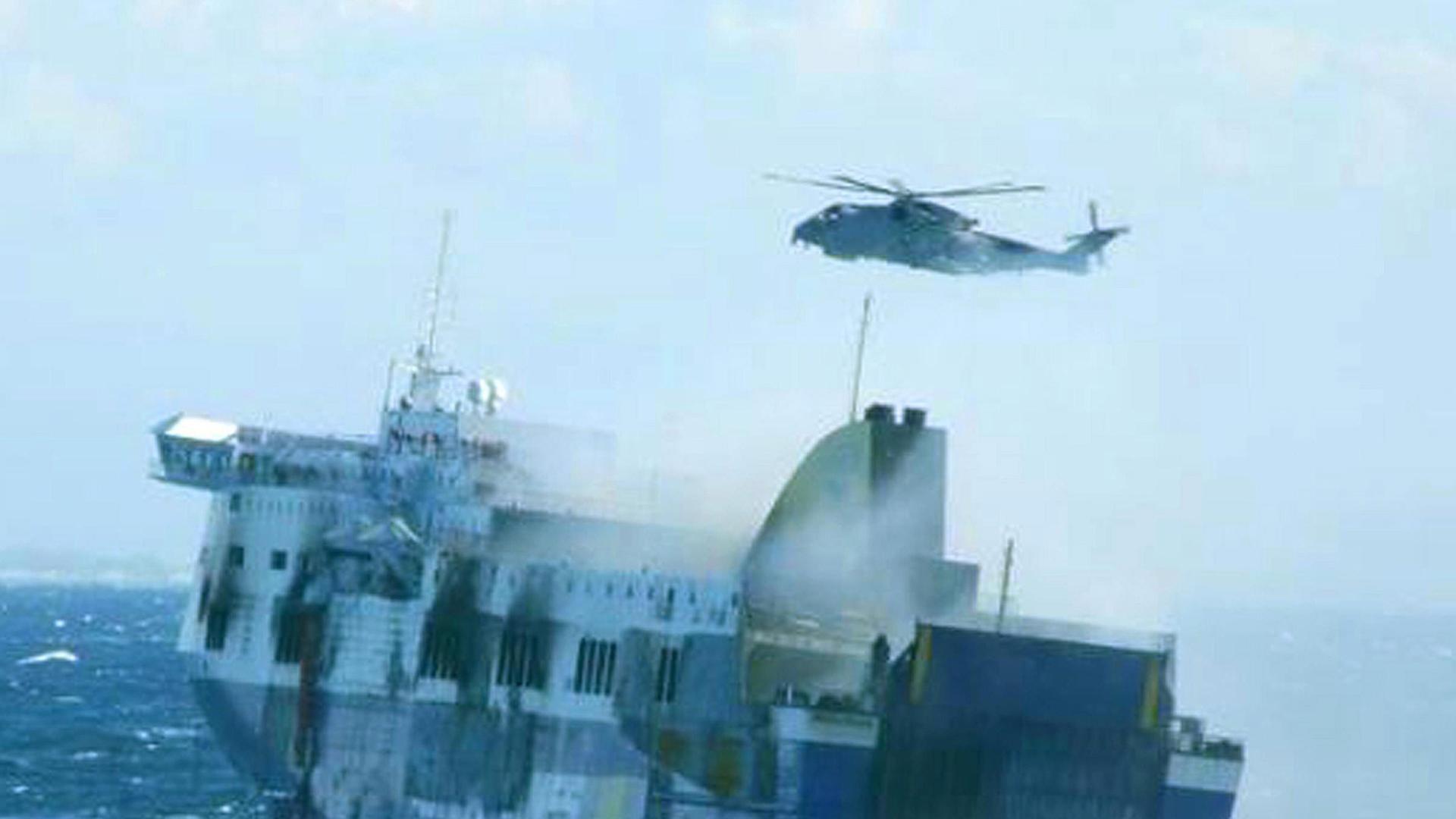 Die Fähre treibt schwankend auf der aufgewühlten See, darüber steht ein Hubschrauber in der Luft.