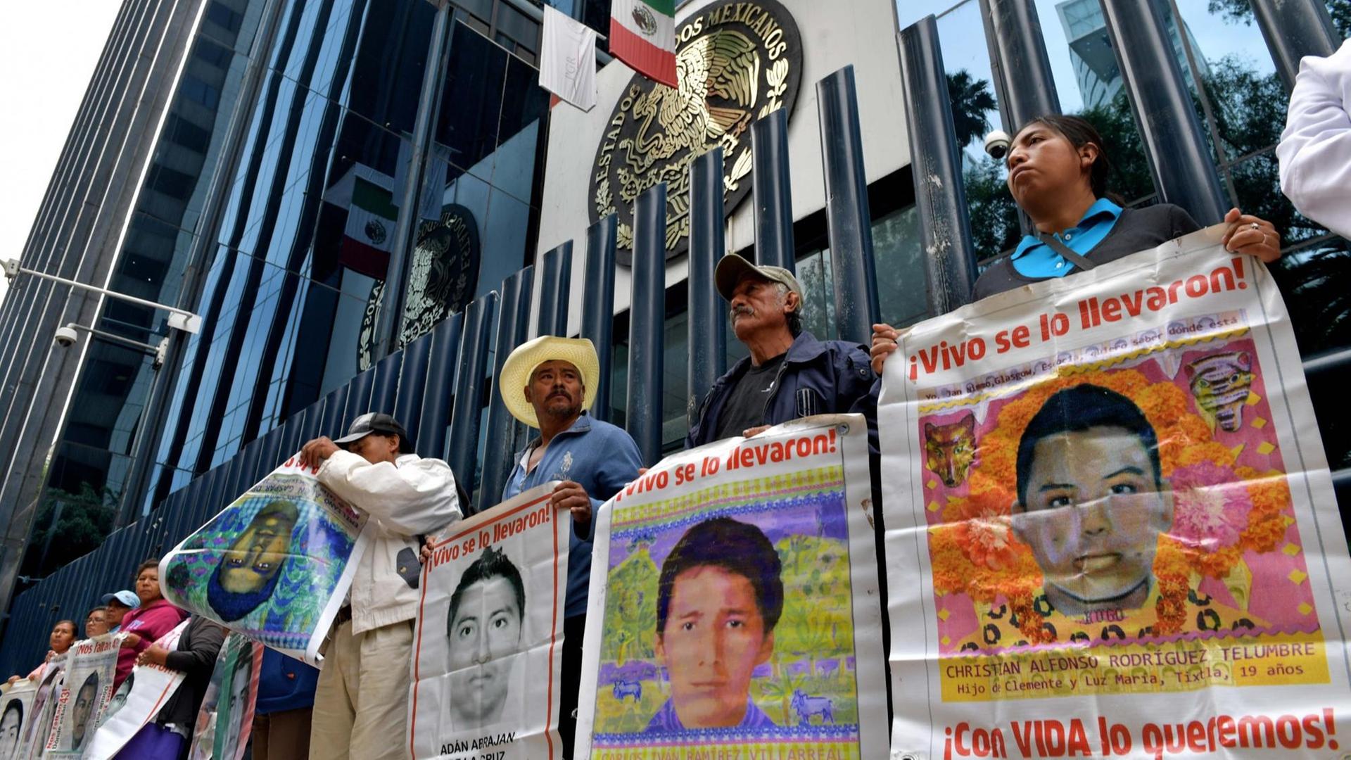 Menschen stehen vor einem Gebäude und halten anklagend Plakate mit Bildern von vermissten Angehörigen und Freunden hoch.