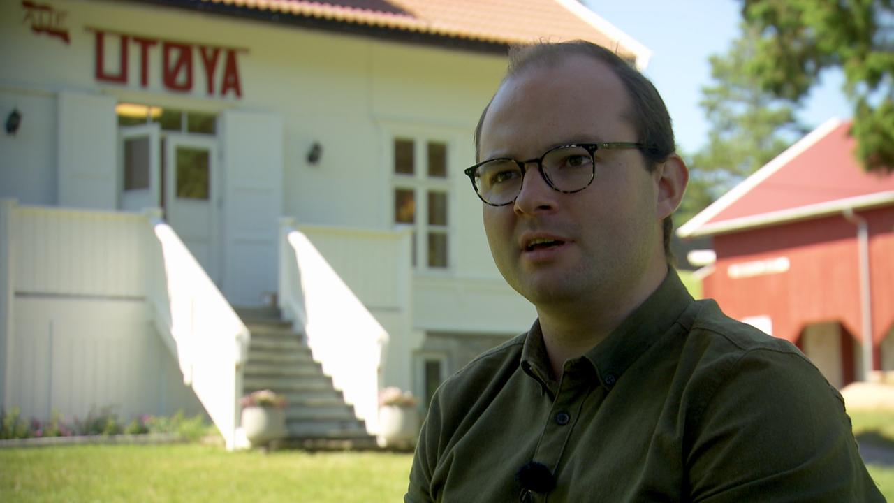 Ein Mann mit Brille, fliehender Stirn und glatt nach hinten gekämmtem Haar steht vor einem weißen Gebäude auf dem mit roten Buchstaben Utøya steht.