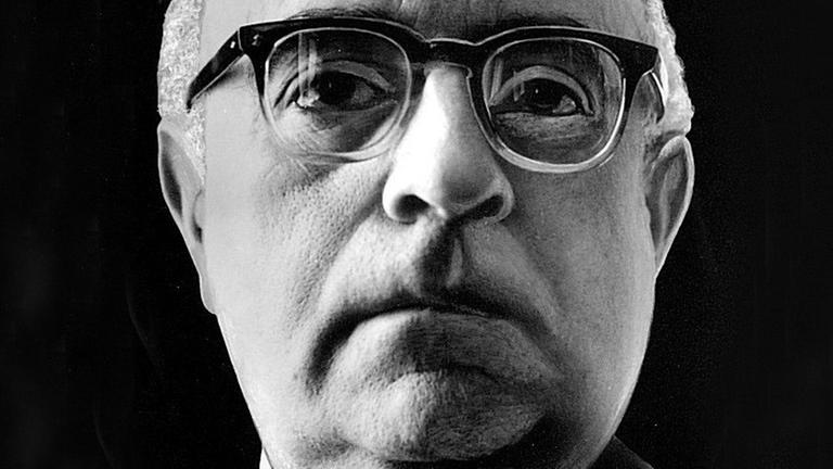 Ein Nahaufnahme von Theodor W. Adorno vor schwarzem Hintergrund. Er trägt eine Hornbrille und schaut aufmerksam dem Betrachter entgegen.