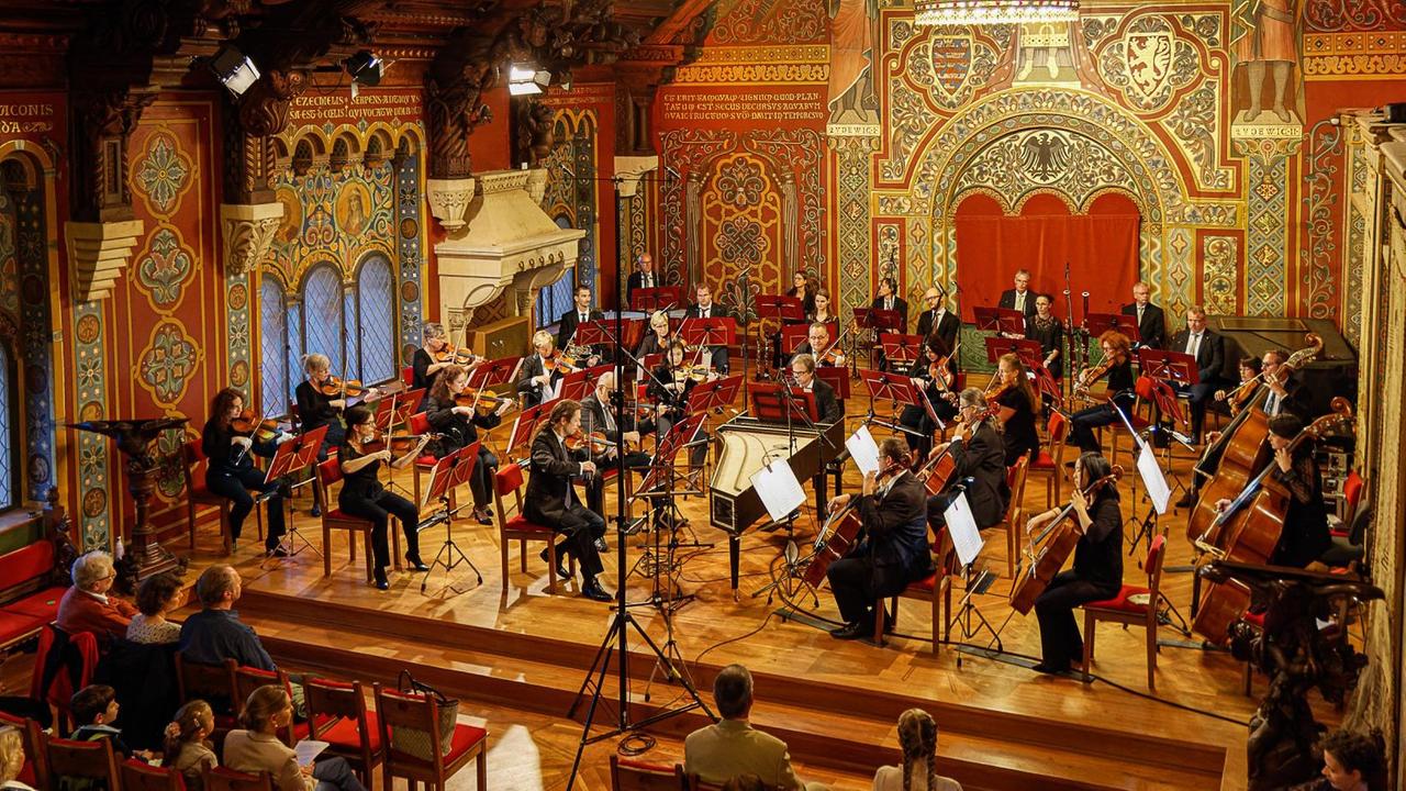Musiker spielen auf einer Bühne im festlichen Palas der Wartburg vor Publikum.