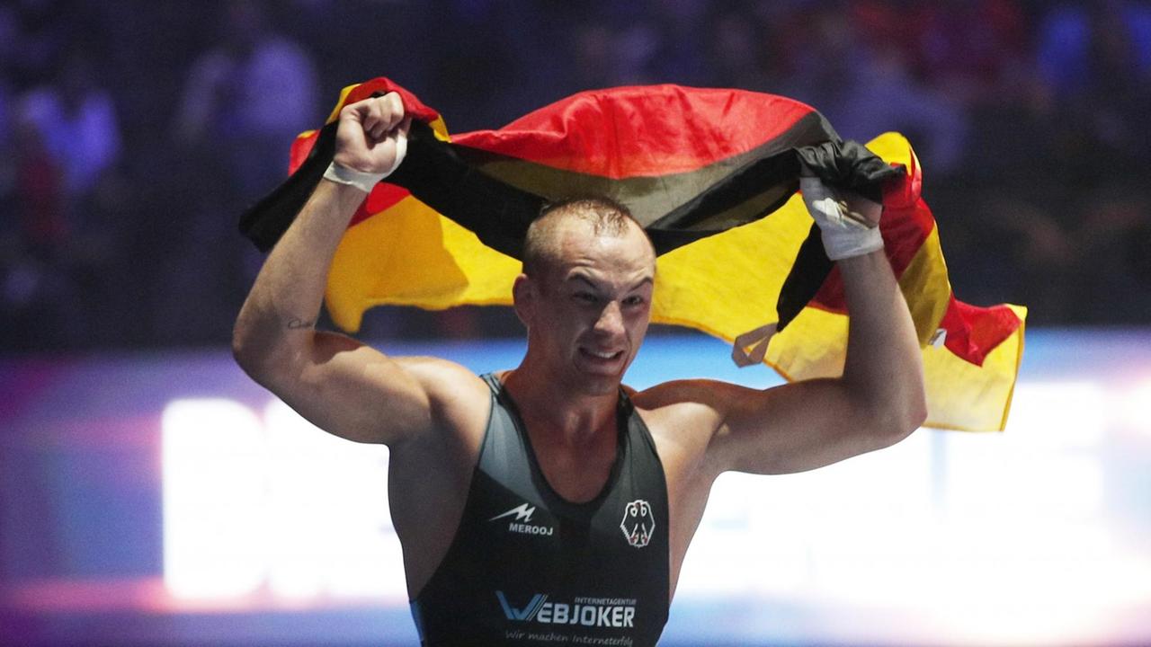 Stäbler im Ringer-Dress hält im Ring eine wehende Deutschland-Fahne über seinen Kopf. Sein Gesicht ist freudig verzerrt.