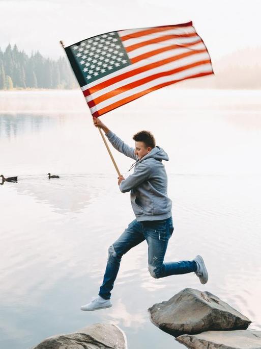 Ein junger Mann springt locker über Steine, die aus einem Gewässer ragen, während er stolz die Flagge der Vereinigten Staaten von Amerika wedelt. Im Hintergrund sieht man im Nebel versunkenes Waldgebirge.