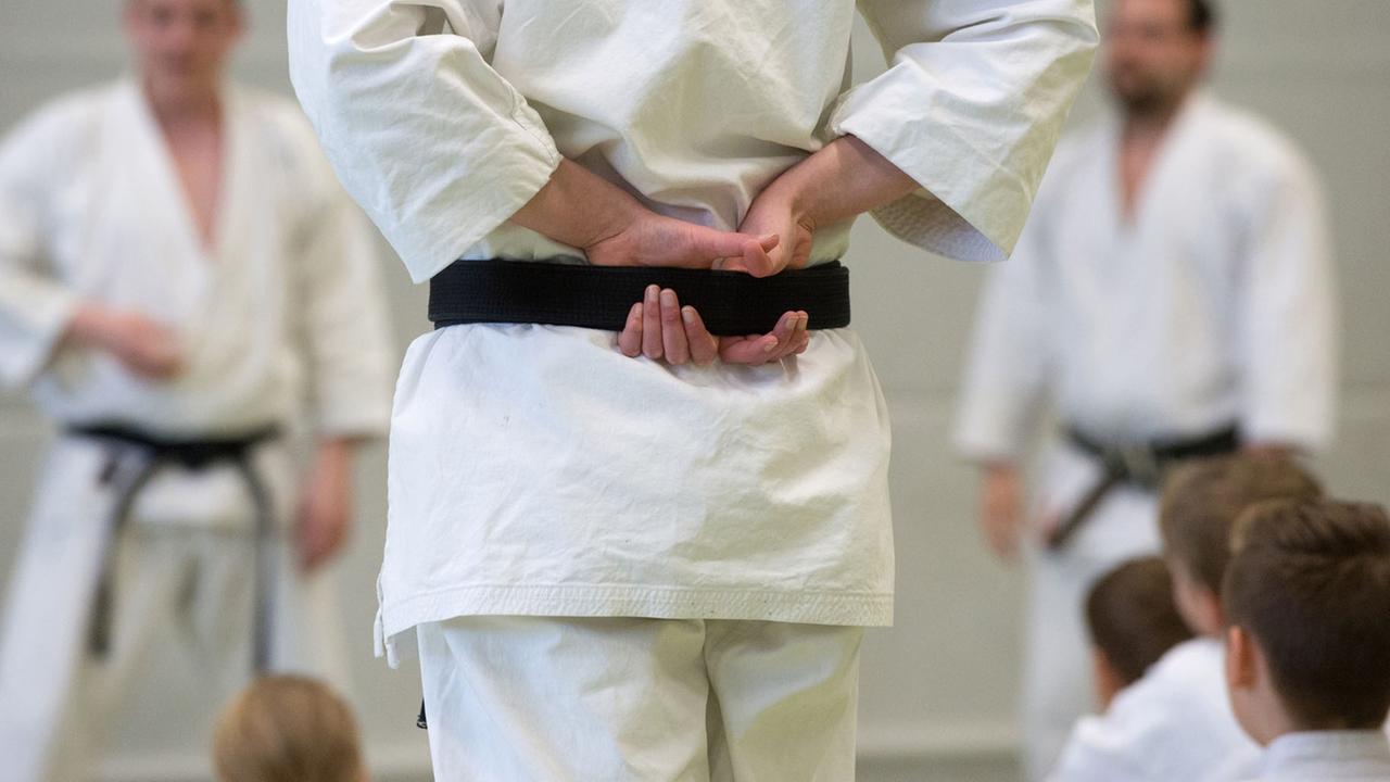 Eine Karate-Trainerin hat ihren schwarzen Gürtel mit beiden Händen auf dem Rücken umfasst, während sie das Training ihrer Schüler kritisch beobachtet.