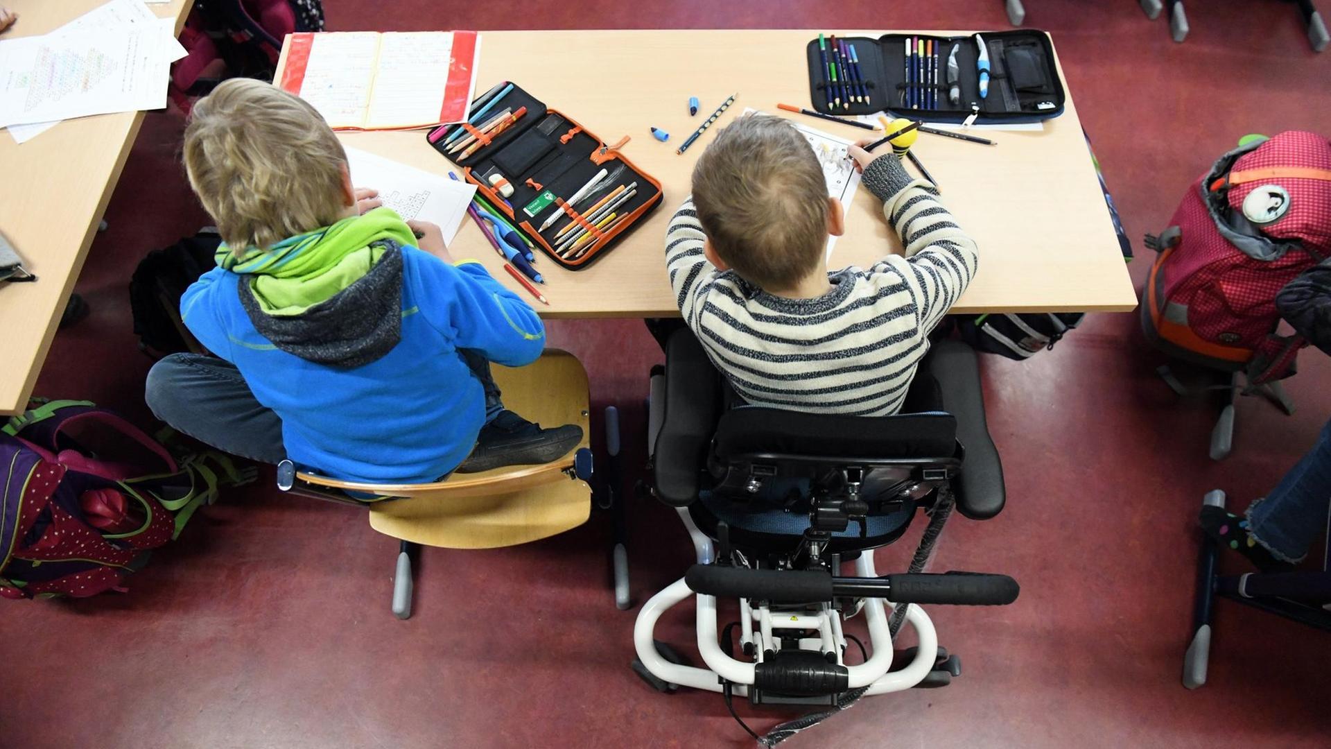 Ein Kind im Rollstuhl sitzt mit einem zweiten Kind an einem Schultisch.