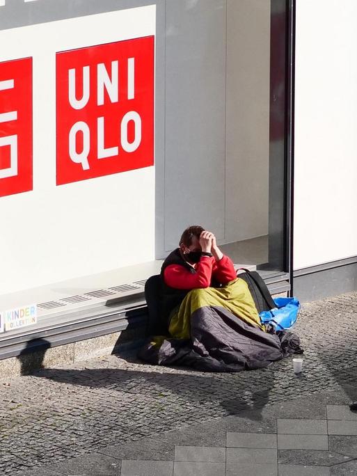 16.05.2020, Berlin. Ein Obdachloser sitzt an einer Einkaufsstrasse in Steglitz vor einem Laden und traegt einen Mundschutz. Ein älterer Mann mit Mundschutz geht an ihm vorbei.