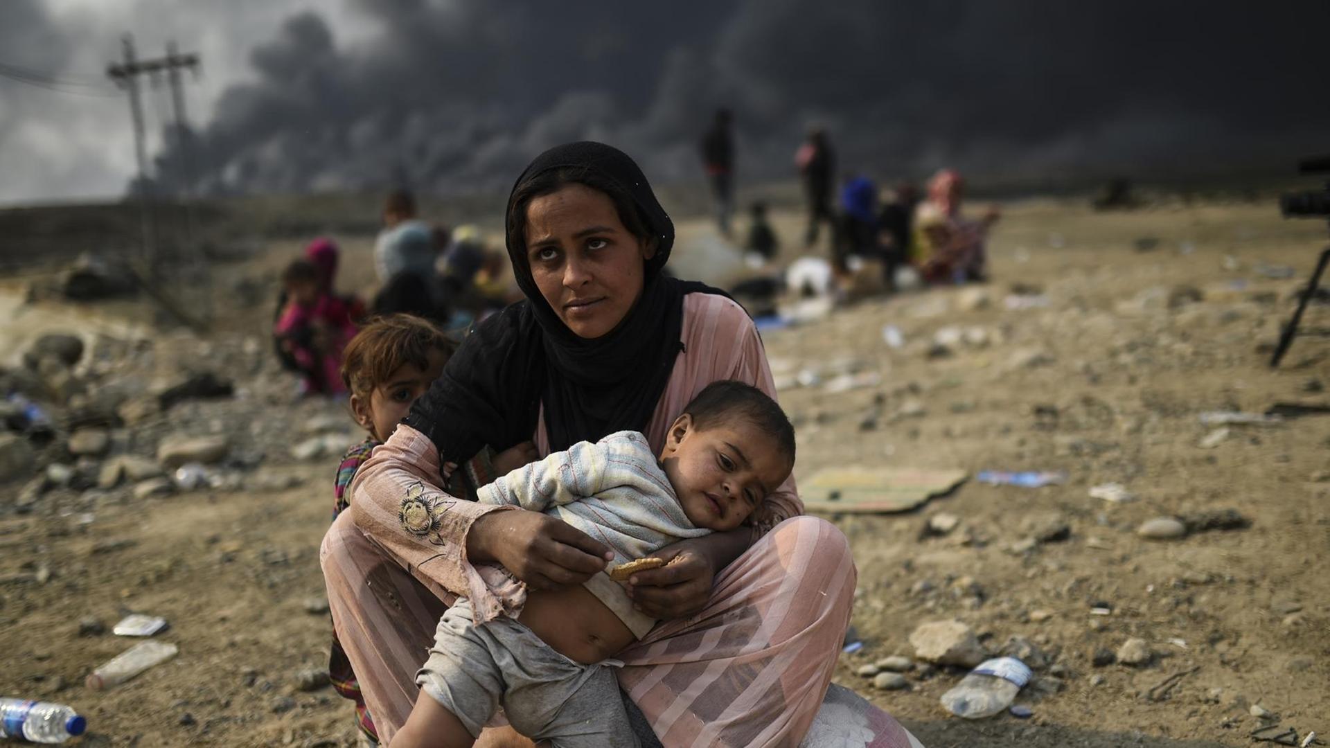 Bild einer Frau mit zwei Kindern, im Hintergrund ist eine schwarze Rauchsäule zu sehen. Aufgenommen in der Region Mossul.
