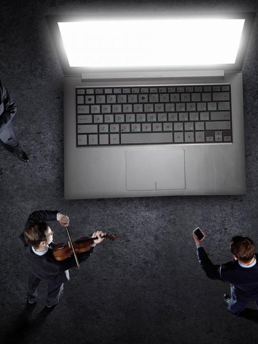 Vier Personen stehen um einen überdimensionierten Laptop und sind dem Bildschirm zugewandt. Einer spielt Geige, die anderen hören mehr oder weniger konzentriert zu.