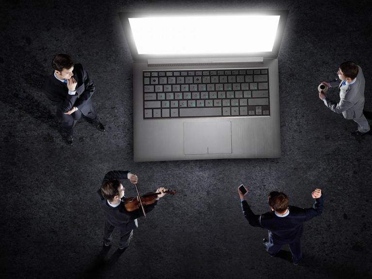 Vier Personen stehen um einen überdimensionierten Laptop und sind dem Bildschirm zugewandt. Einer spielt Geige, die anderen hören mehr oder weniger konzentriert zu.