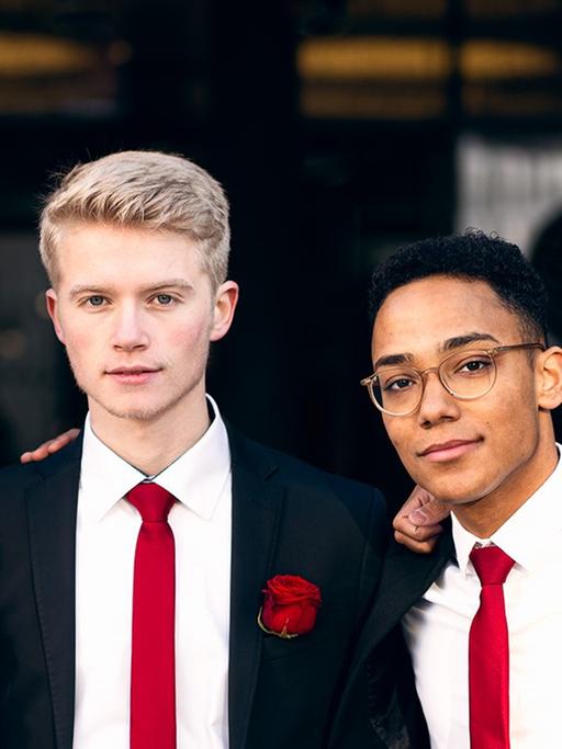 Vier junge Männer in schwarzen Anzügen, roten Schlips und jeweils einer roten Rose am Revers lächeln eng beieinander stehend in die Kamera.