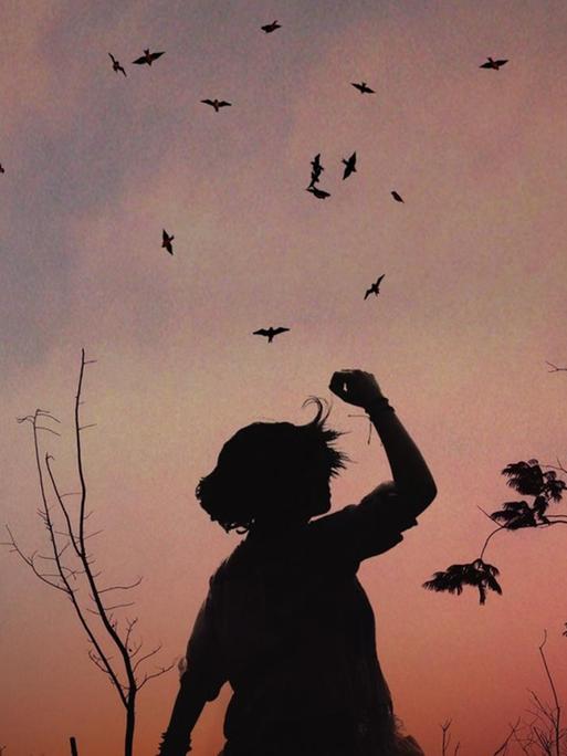 Dämmerungsaufnahme auf einer Lichtung mit den Silhouette eines Mädchens, die ihren Arm in die Höhe hält und einem Vogelschwarm über ihr.