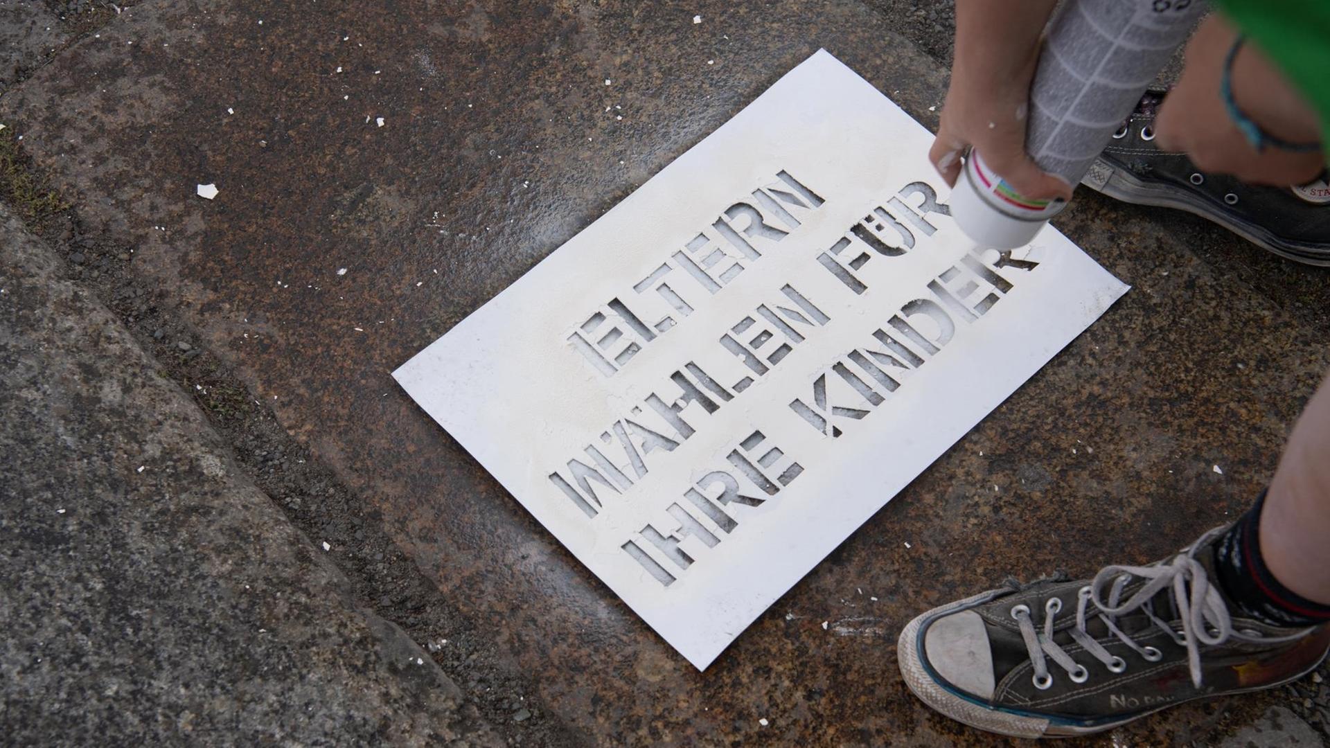 Jemand schreibt mit weißer Farbe und einer Schablone die Worte "Eltern wählen für ihre Kinder" auf den Boden.
