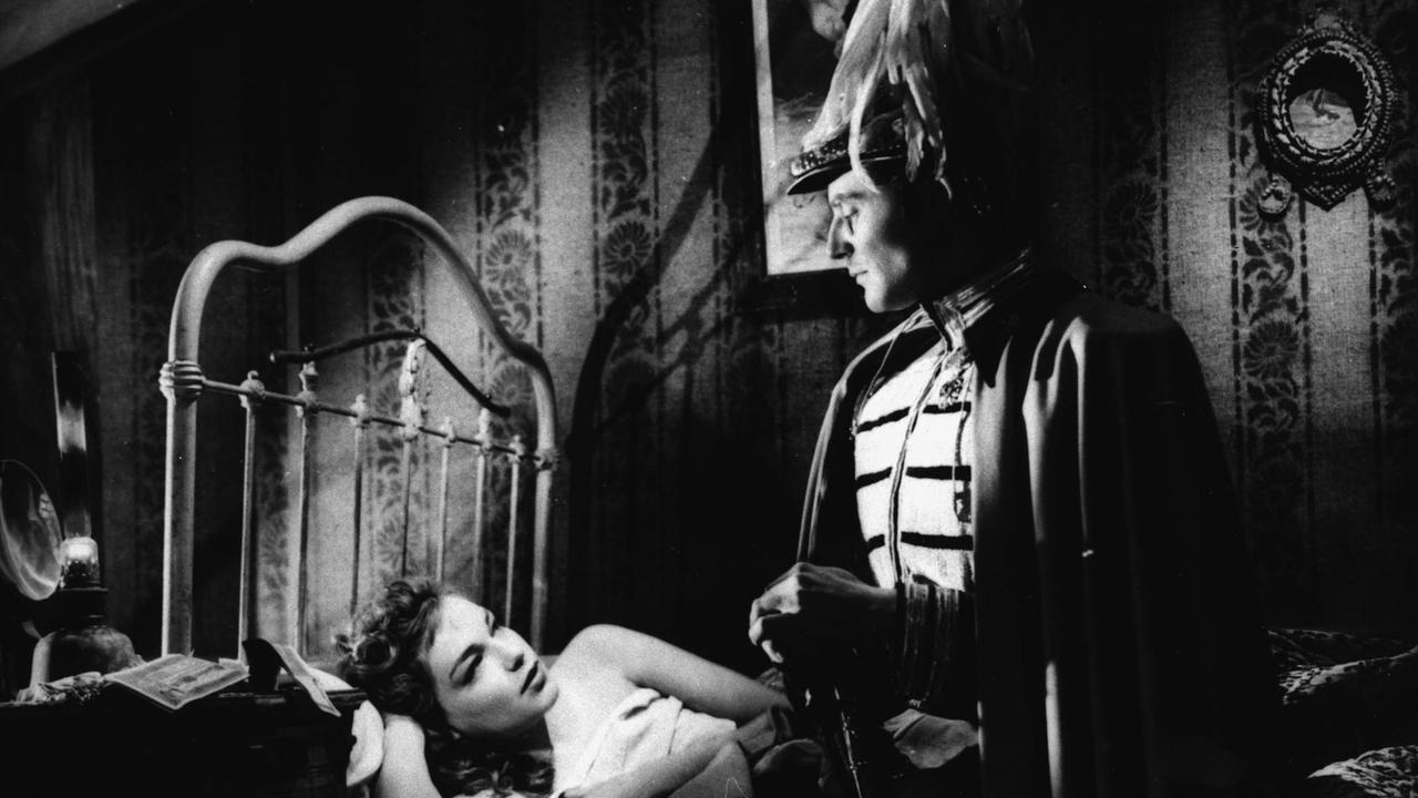 Ein schwarzweißes Filmstill aus Max Ophüls ' Film "Der Reigen" (La Ronde) von 1950 zeigt eine Szene mit Gerard Philippe und Simone Signoret, ihn in Uniform stehend dieauf dem Bett liegende schmachtend anblickend