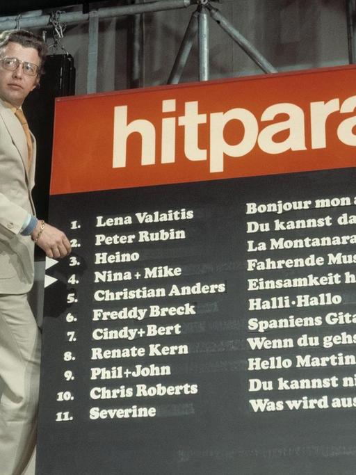 Moderator Dieter Thomas Heck steht auf einer Leiter vor dem Chart der Hitparade im Jahr 1974