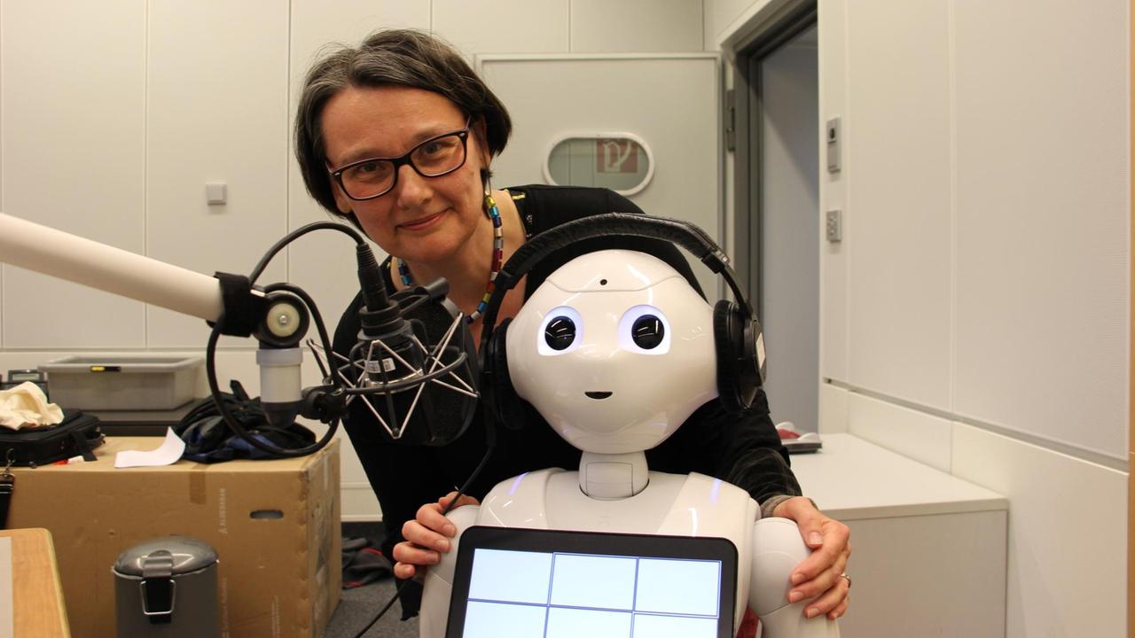 Eine Frau und eine Roboter mit Kopfhörern stehen in einem Radiostudio vor dem Mikrofon.